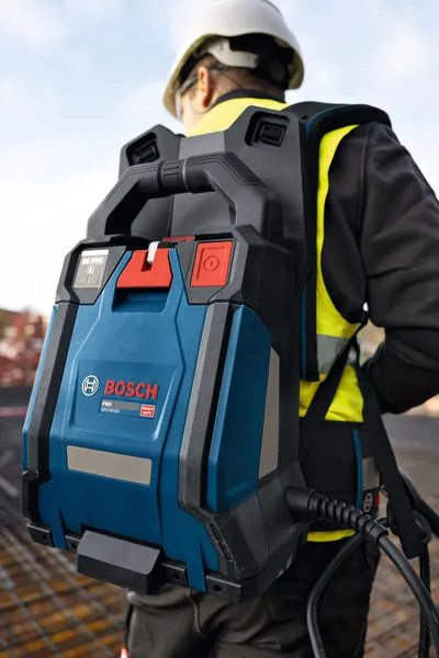 Een persoon met veiligheidsuitrusting draagt ​​een blauwe rugzak met betonvibrator op een bouwplaats.