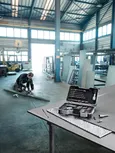 En person med sikkerhetsutstyr måler en metallplate med et bor åpent på et arbeidsbord.