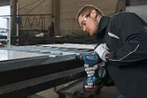 En person med sikkerhetsutstyr borer inn i en metallbjelke i et industrielt miljø.