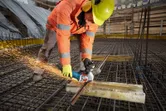 En arbeider iført sikkerhetsutstyr kutter armeringsjern på en byggeplass med en vinkelsliper.