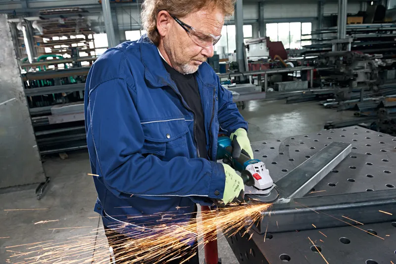 En person med sikkerhetsutstyr sliper en metallbjelke med en vinkelsliper i et verksted.
