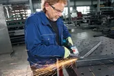 En person med sikkerhetsutstyr sliper en metallbjelke med en vinkelsliper i et verksted.