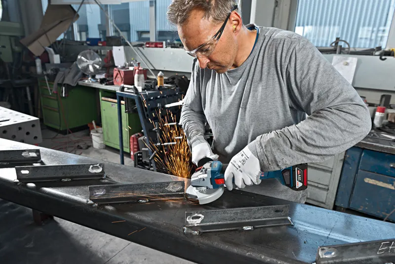 En person med sikkerhetsutstyr sliper metallbjelker med en vinkelsliper, noe som produserer gnister.