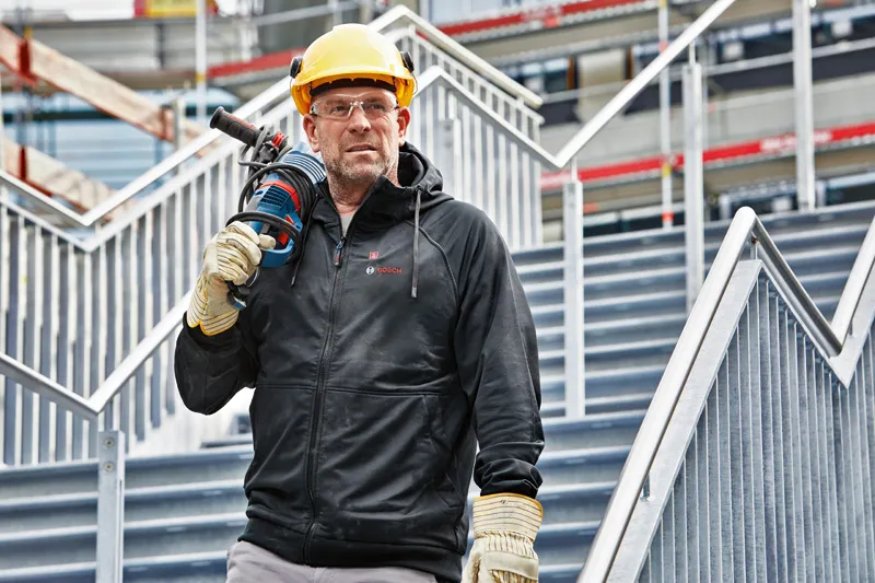 En person med sikkerhetsutstyr og hettegenser med varme står i trappen og holder et elektroverktøy.