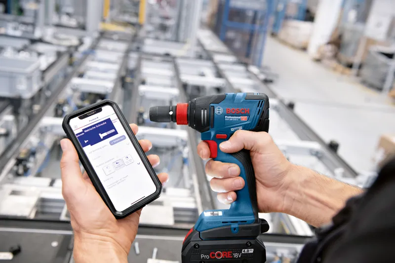 En person bruker en smarttelefon til å styre en trådløs drill i et industrielt miljø.