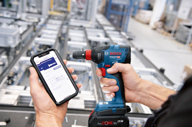 En person bruker en smarttelefon til å styre en trådløs drill i et industrielt miljø.