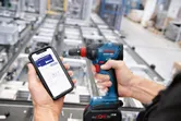 En person bruker en smarttelefon til å styre en trådløs drill i et industrielt miljø.