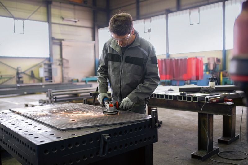En arbeider med sikkerhetsutstyr sliper metall på en stor arbeidsbenk i en fabrikk.