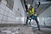 En person med sikkerhetsutstyr bruker en borhammer til å knuse gulvfliser i et flislagt rom.