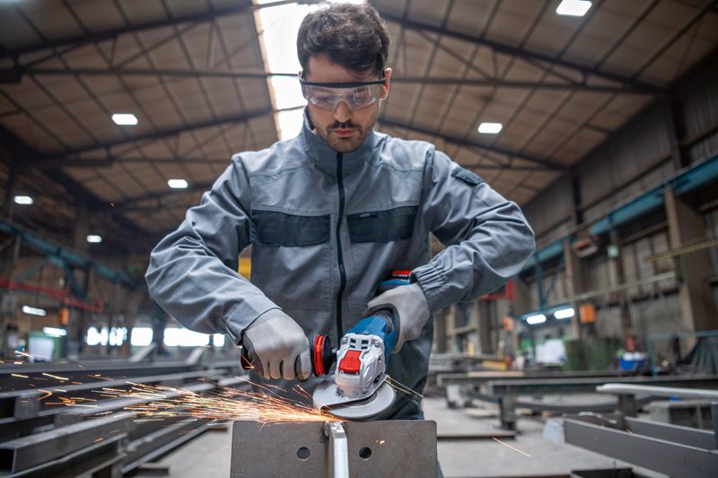 En person med sikkerhetsutstyr sliper metall med en batteridrevet vinkelsliper i en fabrikk.