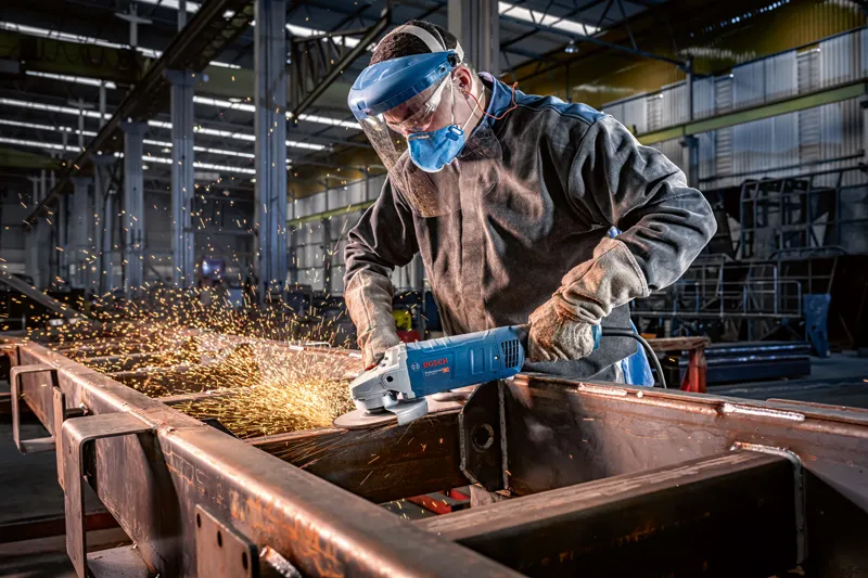 En person med sikkerhetsutstyr sliper metallbjelker med en vinkelsliper, noe som skaper gnister.