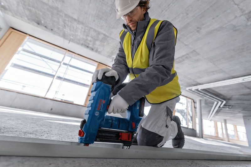En person med sikkerhetsutstyr bruker en batteridrevet betongspikerpistol på en metallskinne.
