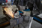 En person med sikkerhetsutstyr sliper en stålbjelke med et elektroverktøy, noe som produserer gnister.