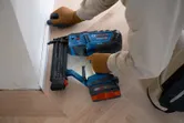 En person som bruker sikkerhetsutstyr bruker en trådløs dykkerpistol til å montere listverk på et gulvhjørne.