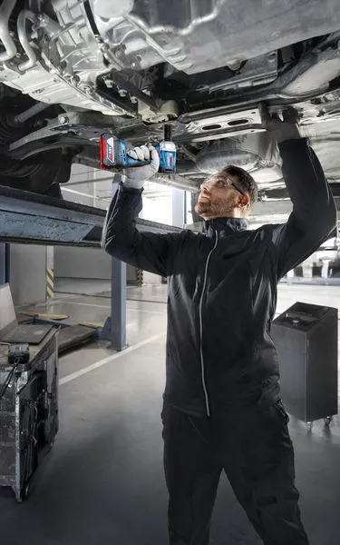 En person med sikkerhetsutstyr bruker en elektrisk drill til å arbeide under et hevet kjøretøy.