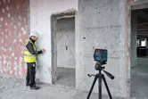 En person med sikkerhetsutstyr markerer en vegg veiledet av et lasernivelleringsverktøy på et stativ.