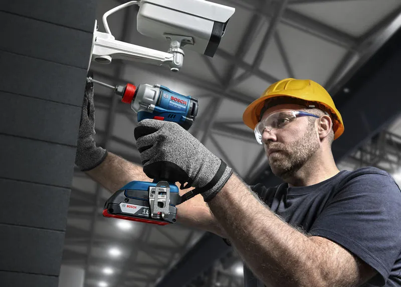 En person med sikkerhetsutstyr bruker en batteridrevet drill til å installere et sikkerhetskamera.