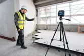 Person med sikkerhetsutstyr måler gulvhøyden med et lasernivelleringsverktøy på et stativ.
