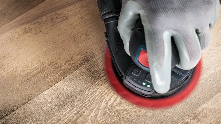 Arbeider iført sikkerhetsutstyr sliper en treflate med en håndholdt slipemaskin.