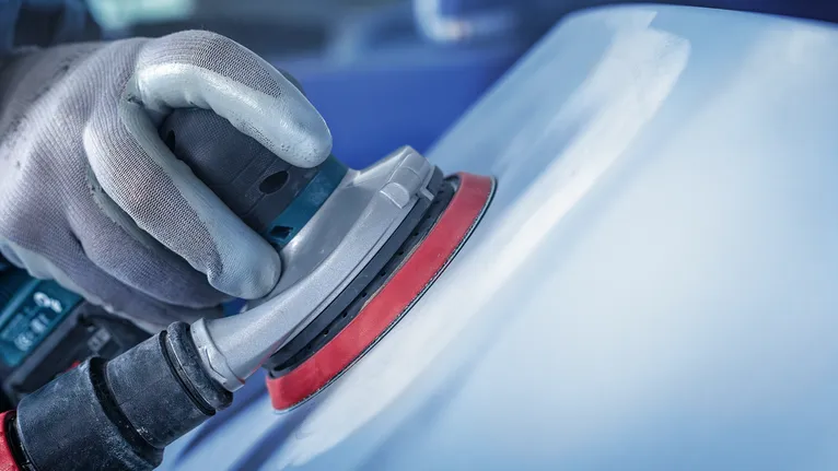 Person med sikkerhetsutstyr sliper en biloverflate med en elektrisk slipemaskin.