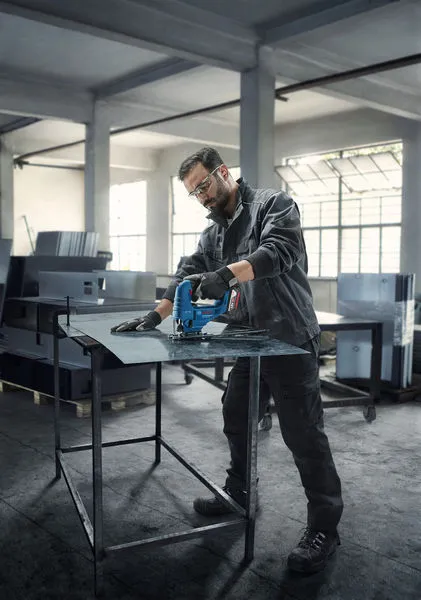 En person med sikkerhetsutstyr skjærer metallplater med et elektroverktøy i et verksted.
