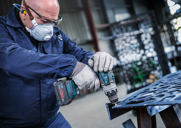 Person med sikkerhetsutstyr borer i en metallplate i et industrielt miljø.