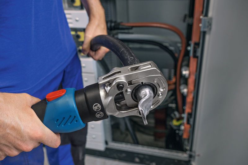 En person bruker et trådløst hydraulisk krympetang på en tykk kabel i nærheten av elektriske paneler.