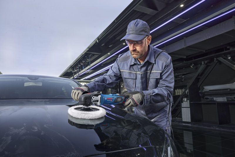 En person med sikkerhetsutstyr polerer en svart bil med en poleringsmaskin.
