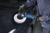 En person med sikkerhetsutstyr bruker en poleringsmaskin på et svart bilpanser.