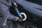 En person med sikkerhetsutstyr polerer en bildør med en elektrisk poleringsmaskin.