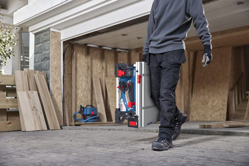 En person med sikkerhetsutstyr bærer en bærbar bordsag på en byggeplass.