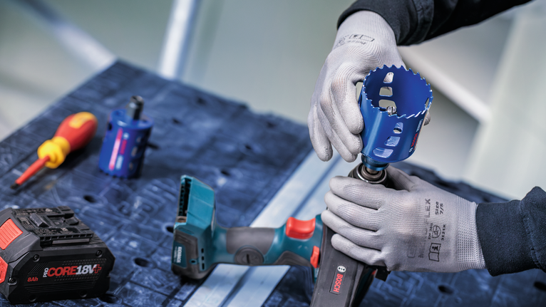 En person med sikkerhetsutstyr fester en hullsag til en batteridrevet drill på en arbeidsbenk.