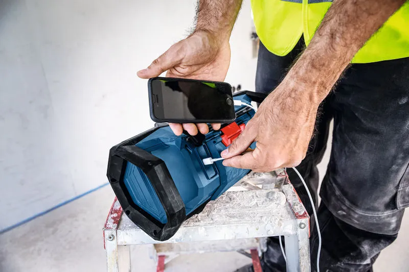 En person med sikkerhetsutstyr lader en smarttelefon ved hjelp av en bærbar arbeidsplassradio.
