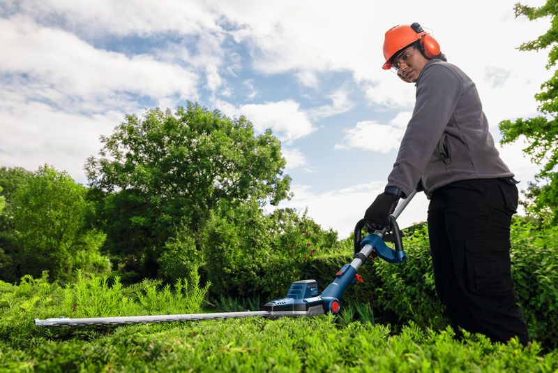 En person med sikkerhetsutstyr trimmer hekker med en batteridrevet hekksaks utendørs.