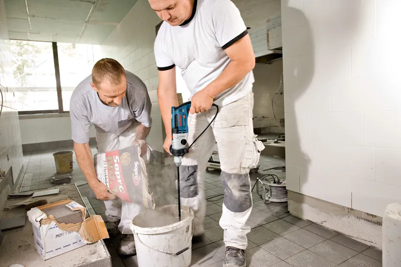 To arbeidere forbereder mørtel, den ene heller pulver mens den andre blander med en elektrisk drill.