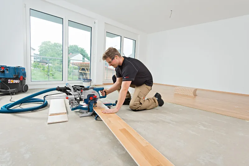 A person wearing safety equipment cuts laminate flooring with a sliding mitre saw.