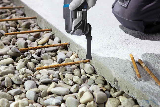 A worker wearing safety equipment uses a tool to cut into concrete near steel rods.