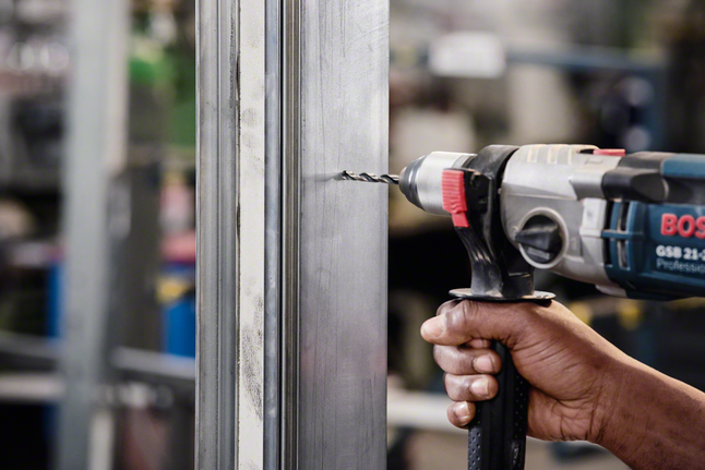 A worker drills a hole through a metal frame indoors.