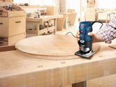 Person guides a palm router along the edge of a round wooden board on a workbench.