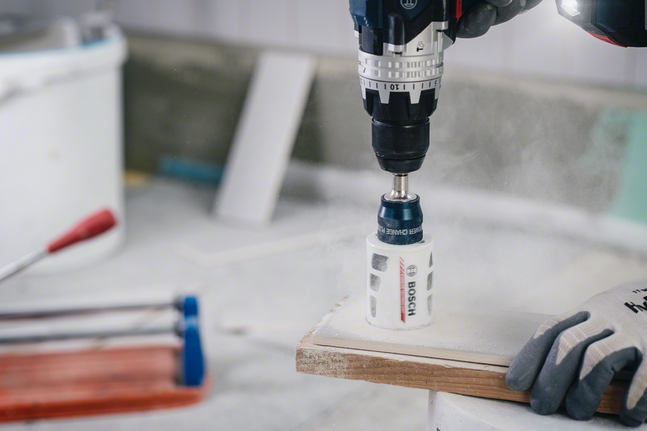 Gloved worker wearing safety equipment drills a hole in tile with a dust-generating hole saw.