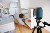 A person installs a cabinet in a kitchen using a laser leveling tool on a tripod.