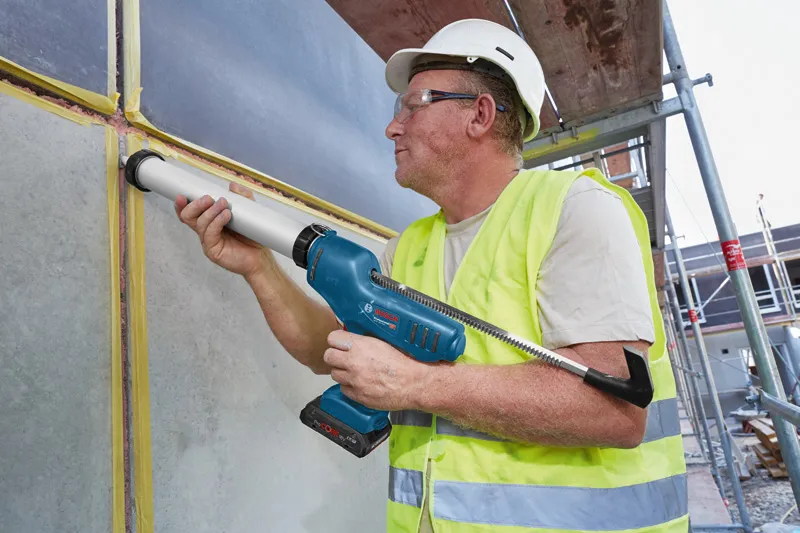 A person wearing safety equipment applies sealant to a wall joint with a cordless caulk gun.