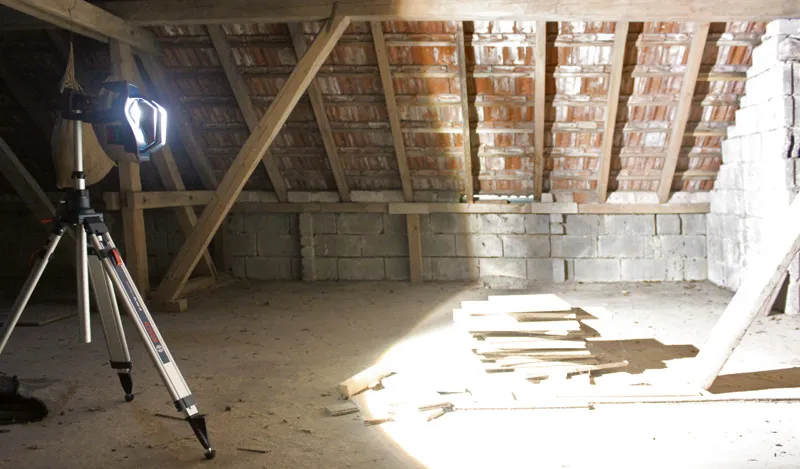 Jobsite light on a tripod brightly illuminates stacked boards in an unfinished attic.