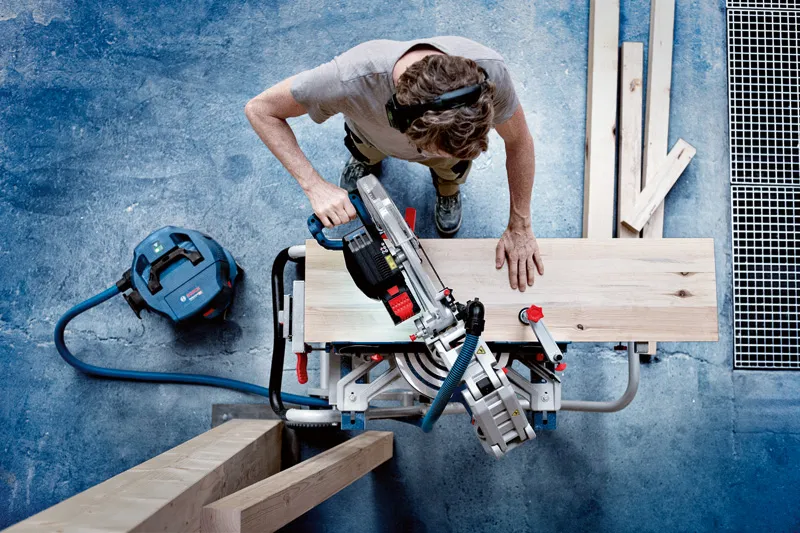 A person wearing safety equipment uses a cordless mitre saw to cut a wooden plank.