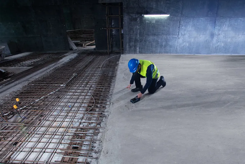 A person wearing safety equipment scans concrete with a detector near exposed rebar.