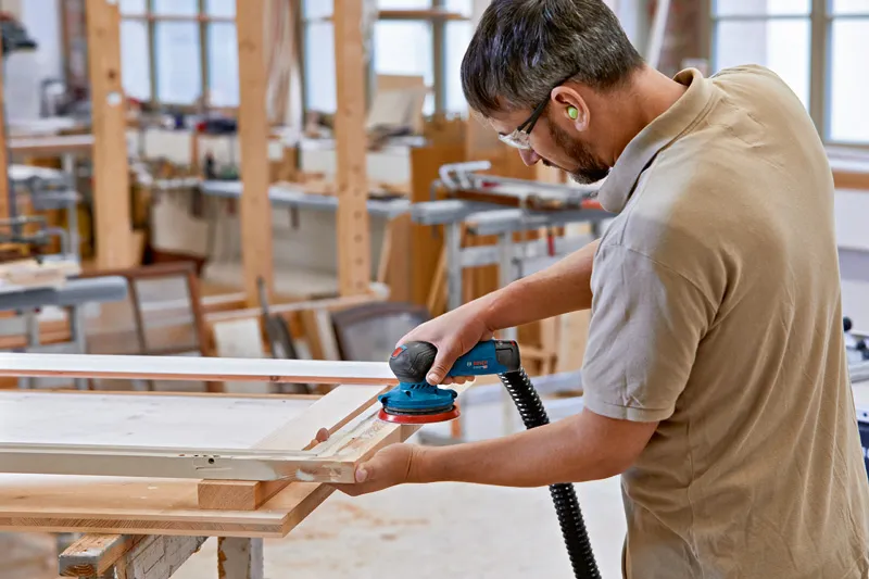 GEX 12V-125 A person wearing safety equipment sands a wooden frame with a cordless random orbit sander.