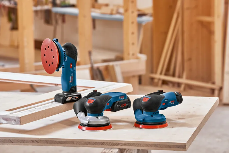 GEX 12V-125 Three electric sanders rest on a wooden workbench in a carpentry workshop.