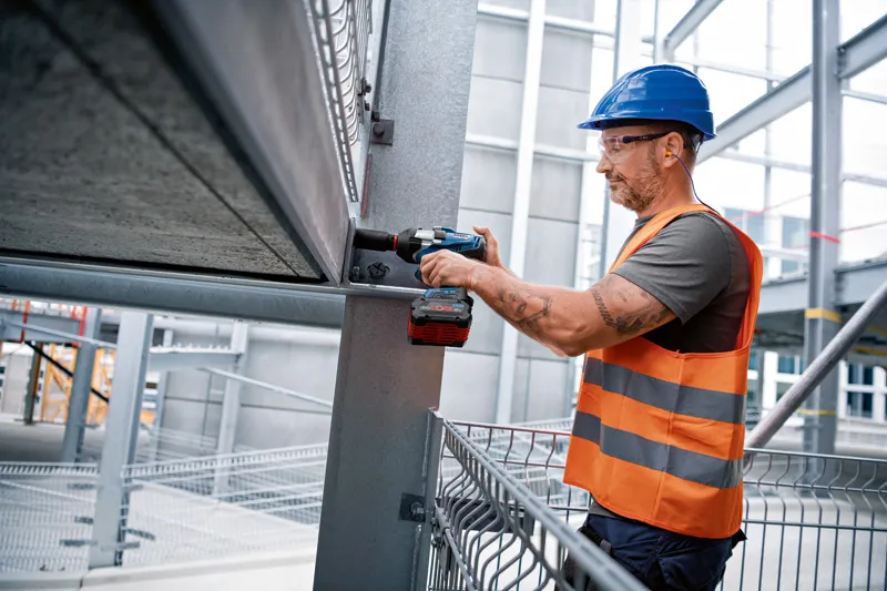 A person wearing safety equipment uses a cordless impact wrench on a steel beam.
