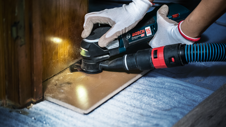 Person wearing safety equipment uses a power tool to sand wood flooring near a cabinet.