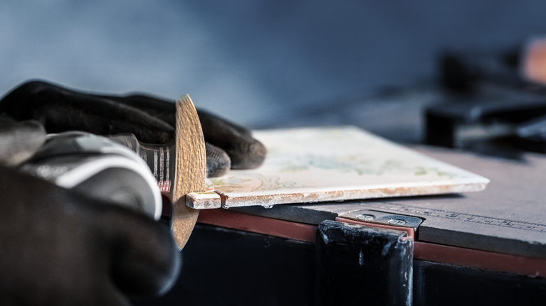 Person wearing safety equipment grinds the edge of a ceramic tile.